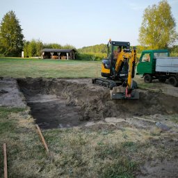 MI-KOP Usługi Ziemne Michał Kuryś - Żółta mini koparka w trakcie prac ziemnych na działce, wykop pod szambo lub oczyszczalnię. W tle altana i zielony krajobraz.