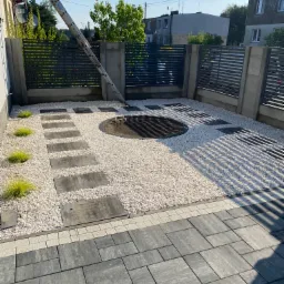 Nowoczesny ogr&oacute;d: ścieżka z płyt, biały żwir, rabata w kształcie koła, metalowe ogrodzenie. W tle budynki mieszkalne i drabina oparta o ścianę.