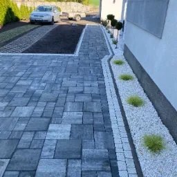 Ułożona szara kostka brukowa prowadzi do domu. Wzdłuż ścieżki rabata z białym kamieniem i ozdobnymi trawami. W tle auto na podjeździe z trawy i kostki.