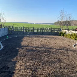 Przygotowany teren pod trawnik, obrzeża z kostki, widok na zielone pole w tle, ogrodzenie panelowe i betonowe. Prace ziemne, wyrównany grunt.