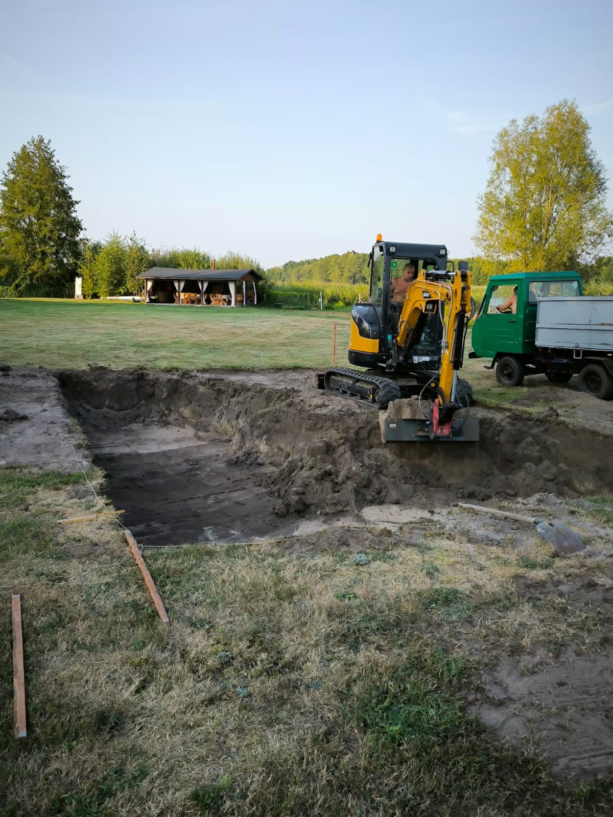 Żółta mini koparka w trakcie prac ziemnych na działce, wykop pod szambo lub oczyszczalnię. W tle altana i zielony krajobraz.