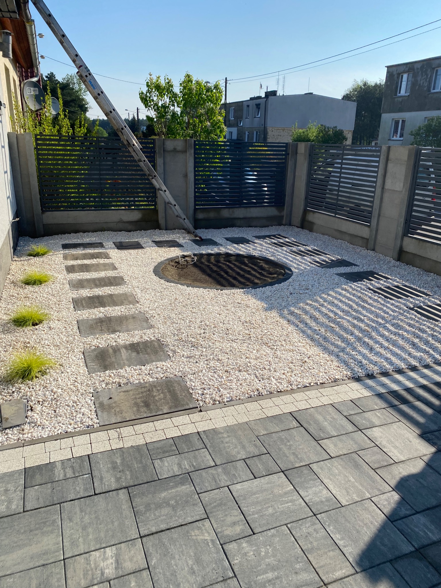 Nowoczesny ogród: ścieżka z płyt, biały żwir, rabata w kształcie koła, metalowe ogrodzenie. W tle budynki mieszkalne i drabina oparta o ścianę.