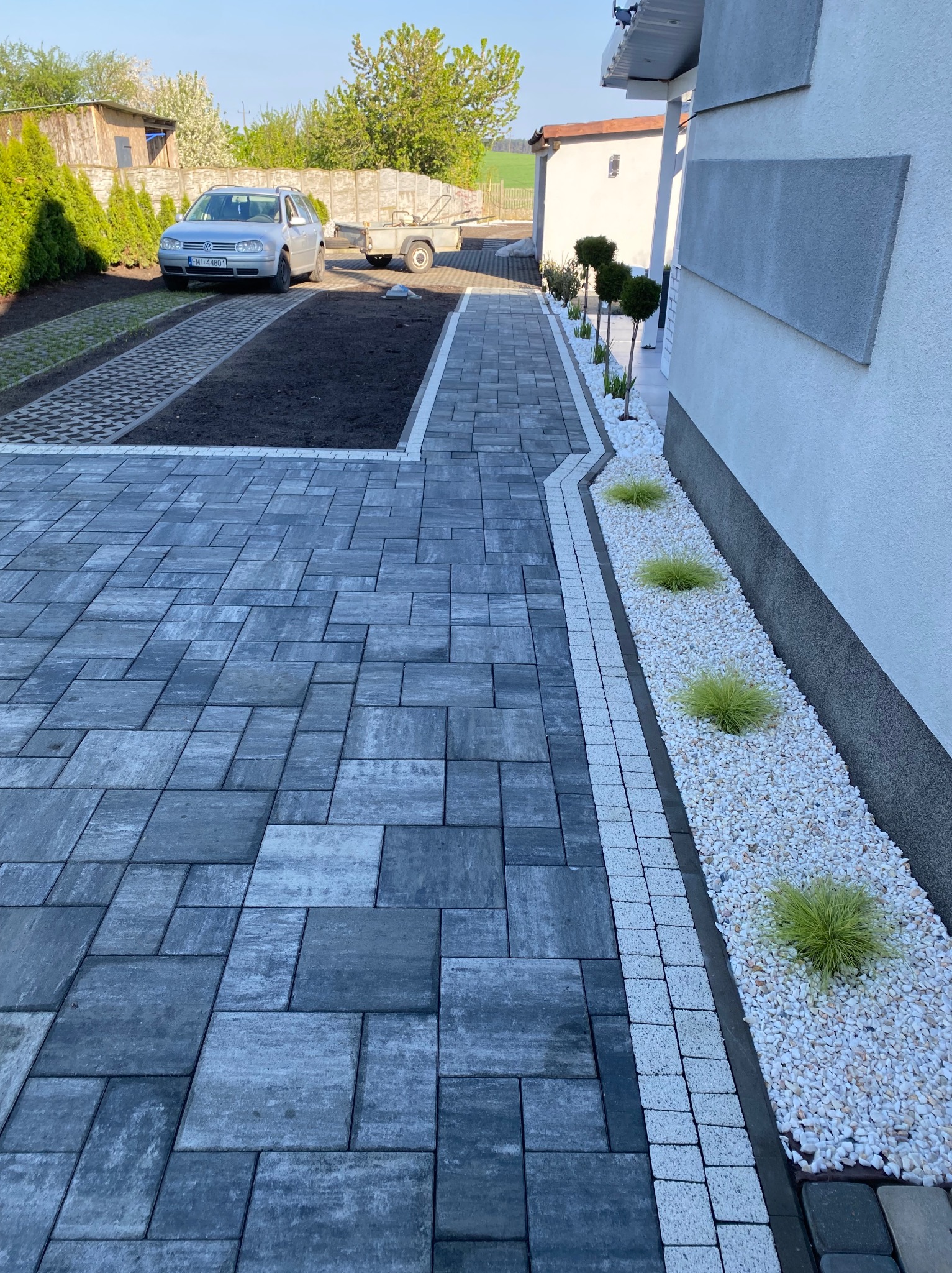 Ułożona szara kostka brukowa prowadzi do domu. Wzdłuż ścieżki rabata z białym kamieniem i ozdobnymi trawami. W tle auto na podjeździe z trawy i kostki.