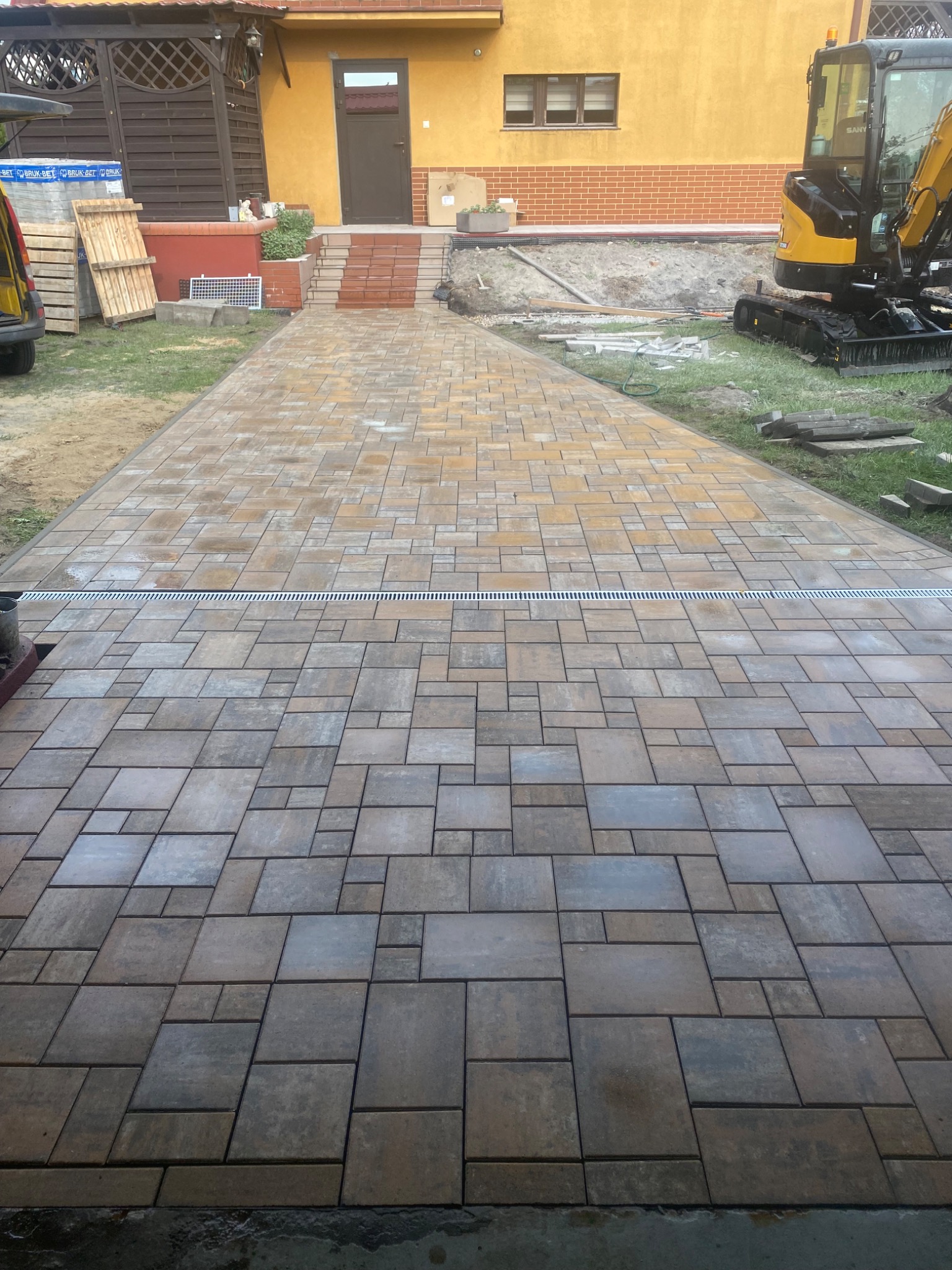 Ułożona kostka brukowa prowadząca do domu z żółtą elewacją. Widoczny drenaż liniowy i mini koparka. Kostka w odcieniach brązu i szarości, ułożona w nieregularny wzór.