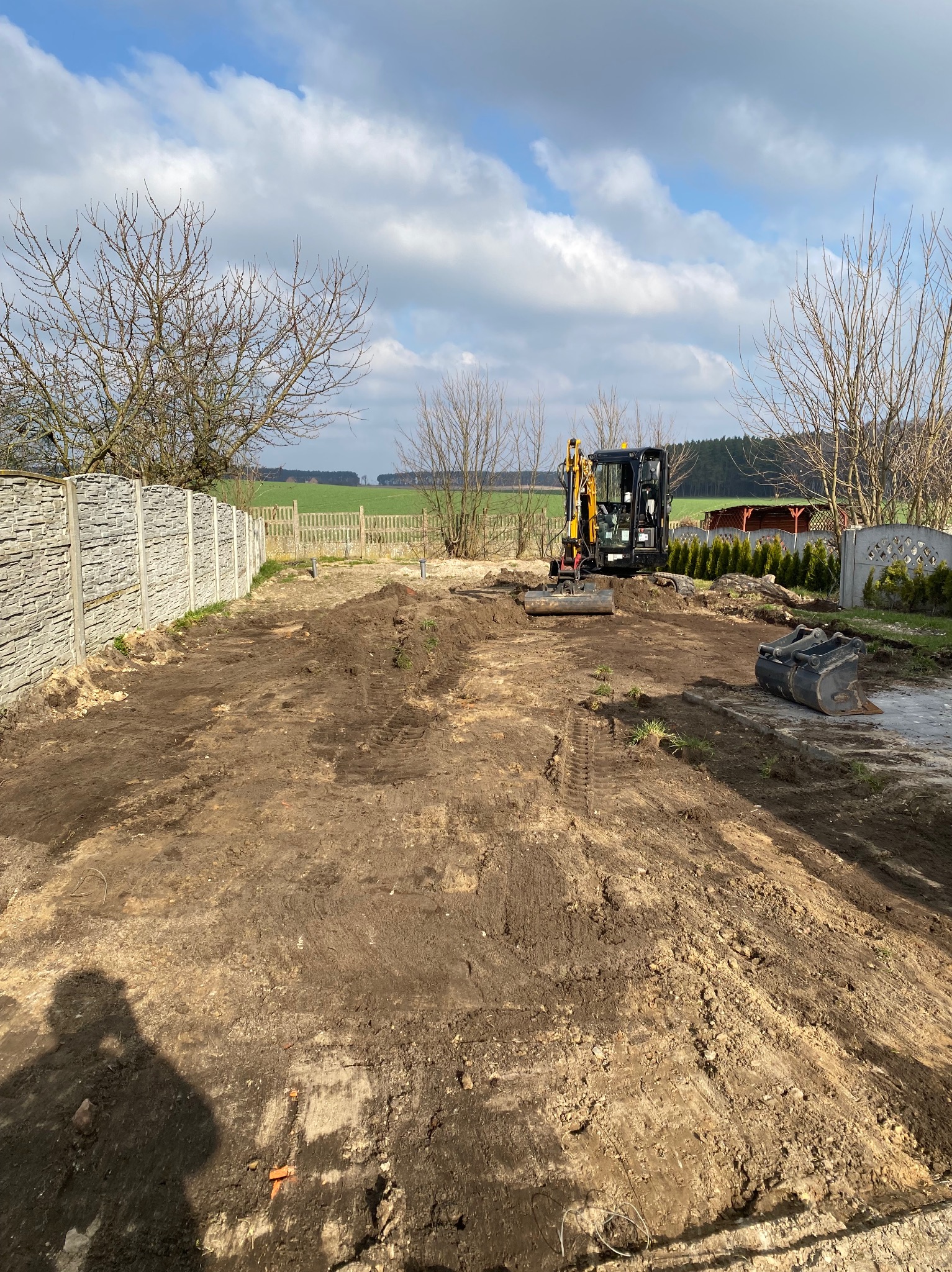 Mini koparka na działce z przygotowanym terenem pod budowę, widoczne ślady opon, ogrodzenie panelowe i shadow operatora w rogu kadru.
