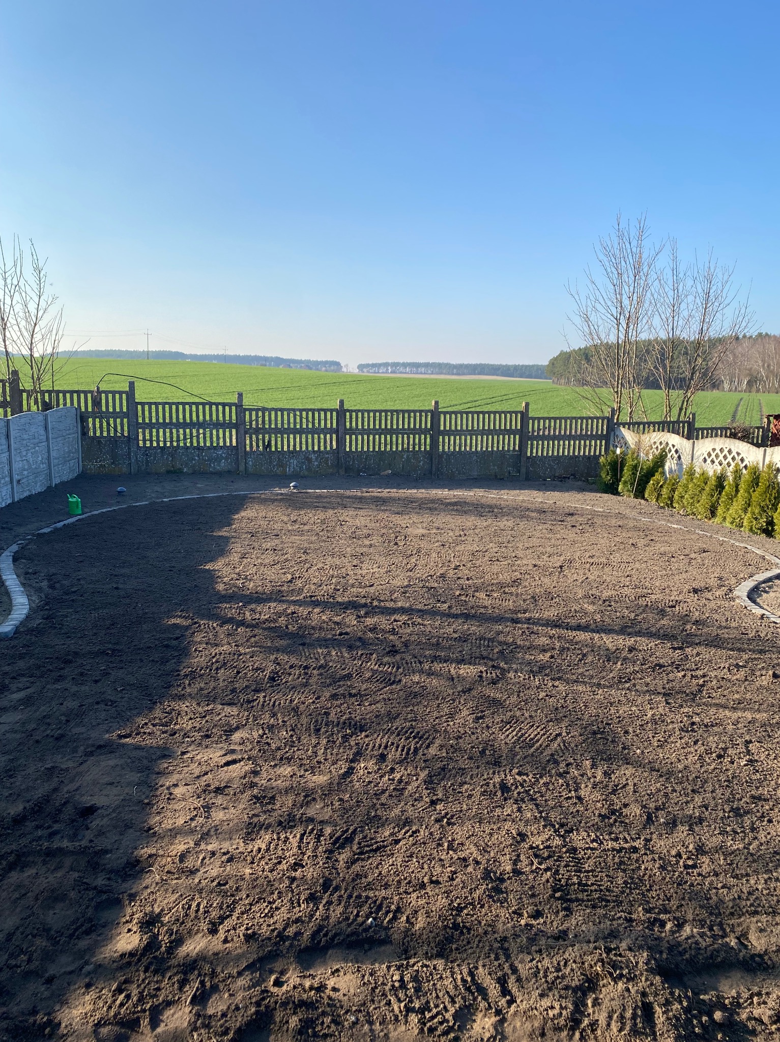 Przygotowany teren pod trawnik, obrzeża z kostki, widok na zielone pole w tle, ogrodzenie panelowe i betonowe. Prace ziemne, wyrównany grunt.