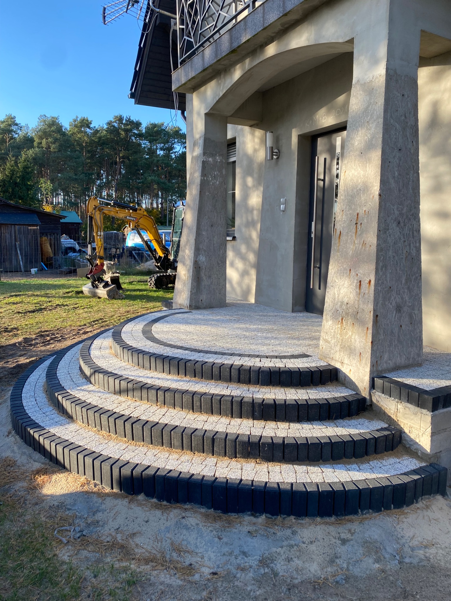 Półkoliste schody z kostki brukowej prowadzące do wejścia budynku. W tle widoczny mały, żółty koparka. Elementy kostki w odcieniach szarości i czerni.