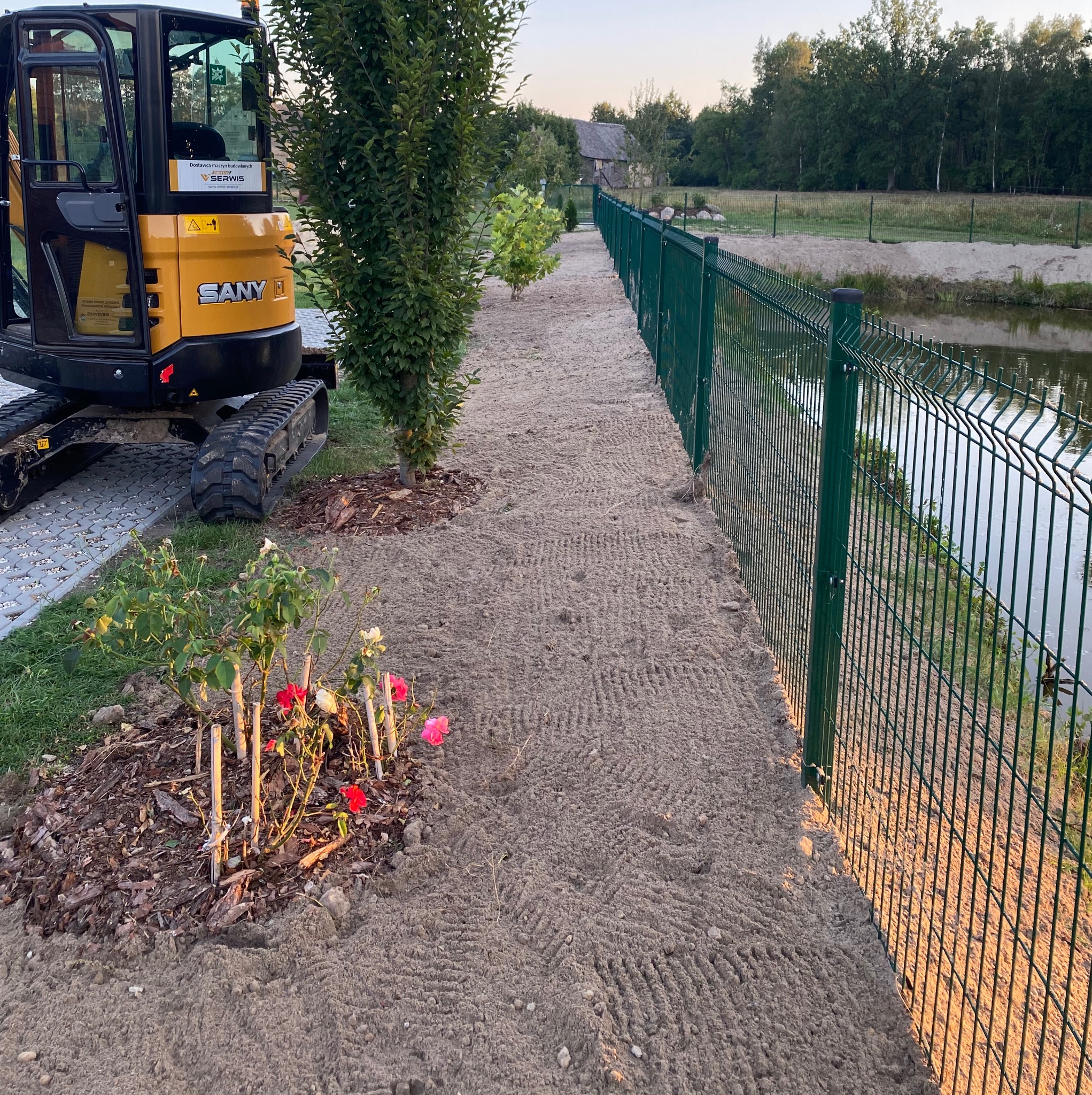 Mini koparka SANY obok zielonego ogrodzenia panelowego, świeżo wyrównany teren, młode drzewka i róże. Widok na staw i budynek w tle.