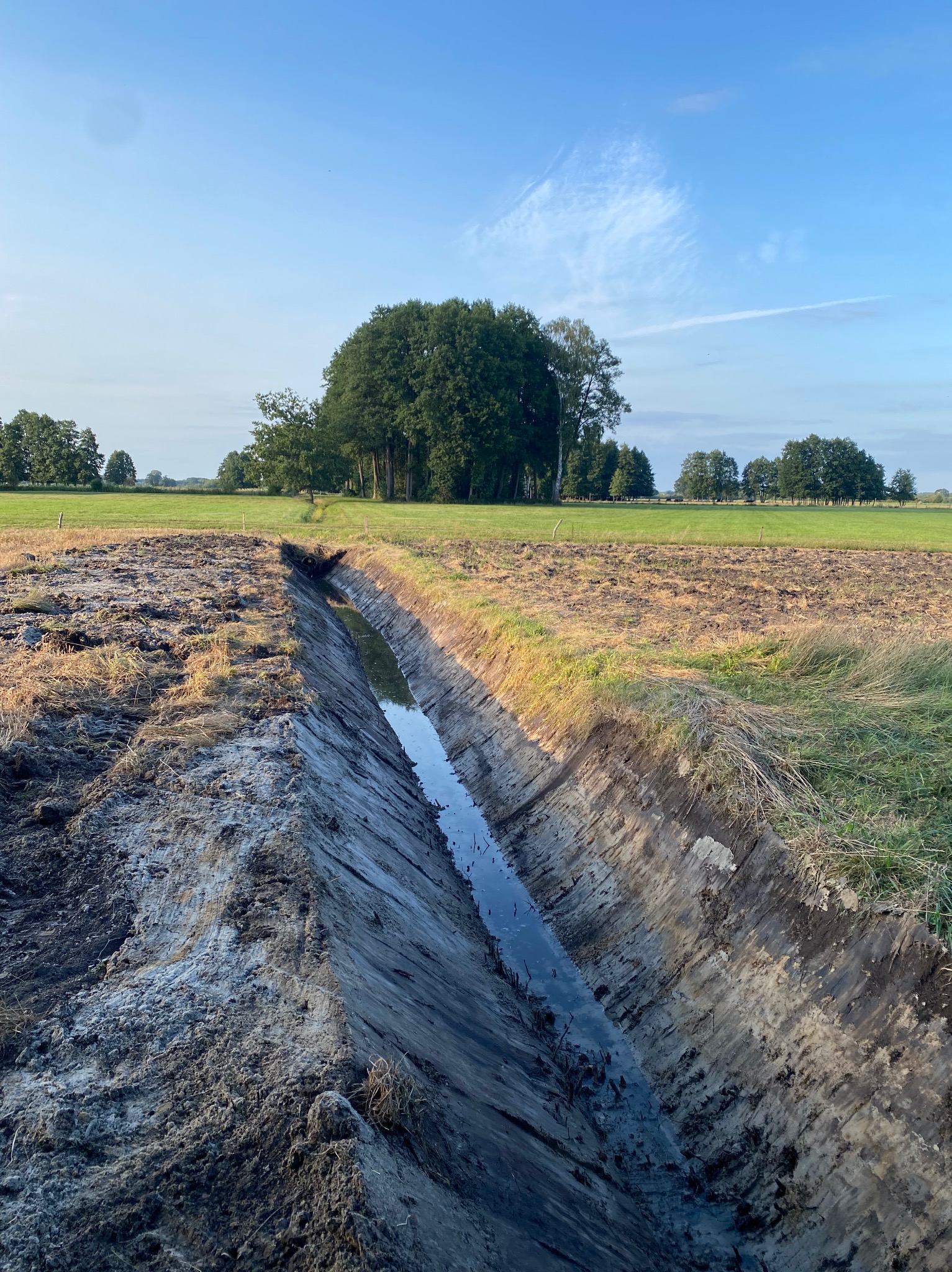 Wykopany rów melioracyjny wypełniony wodą na tle zielonego pola i drzew w oddali, wykonany w słoneczny dzień. Widoczna świeżo przekopana ziemia.