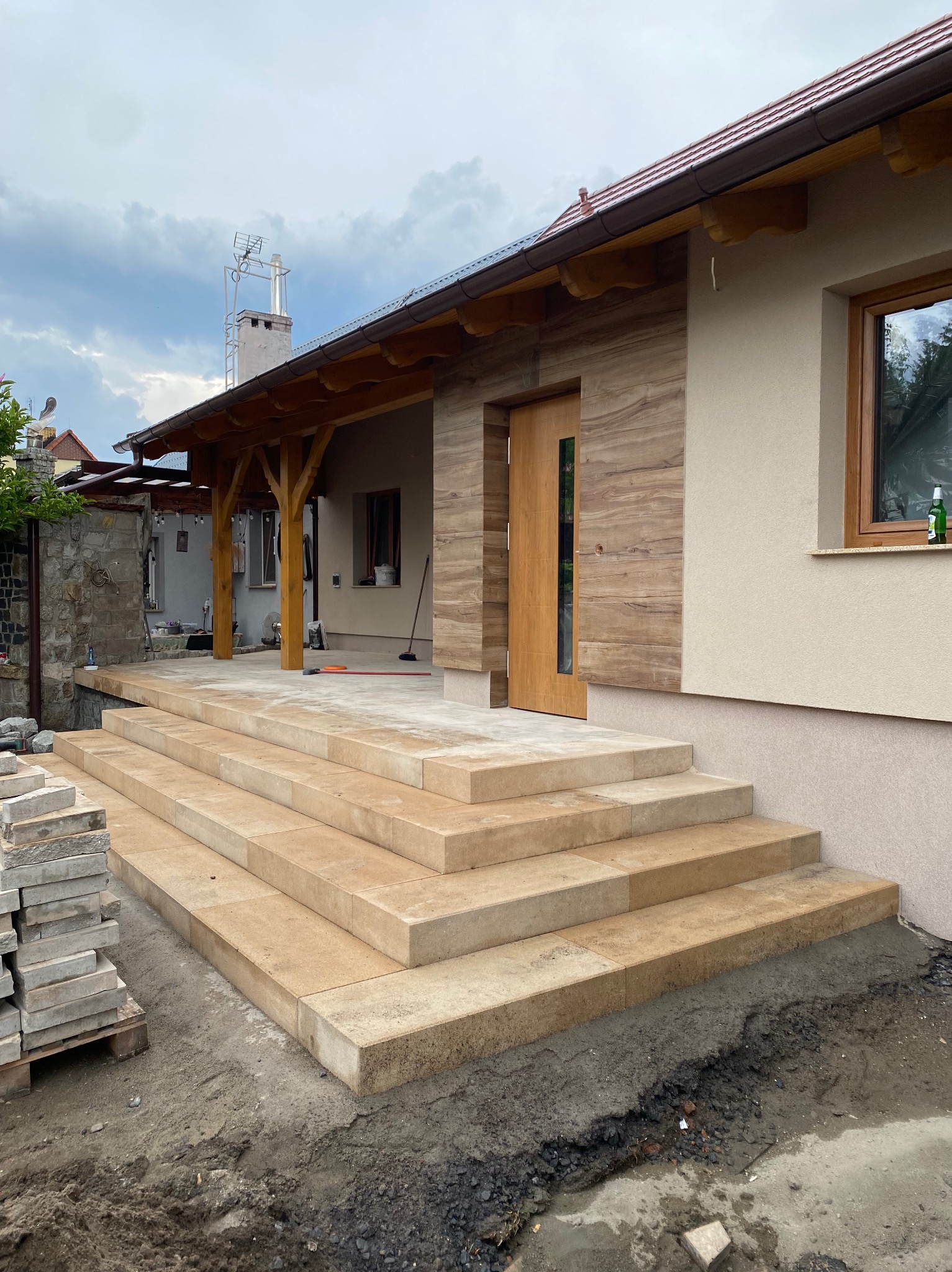 Wykończone schody wejściowe z jasnej kostki brukowej prowadzące do domu z drewnianą elewacją i zadaszeniem. Ułożona kostka na tarasie. Stos kostki brukowej obok schodów.