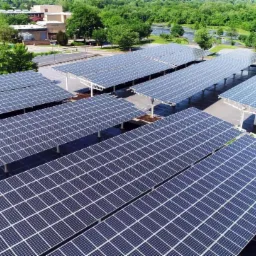 Widok z lotu ptaka na parking z zadaszeniem z paneli słonecznych, budynek w tle, otoczone drzewami.