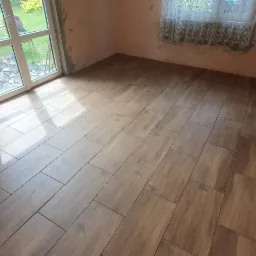 Ułożona podłoga z płytek imitujących drewno w pokoju z surowymi ścianami i oknem na ogród.