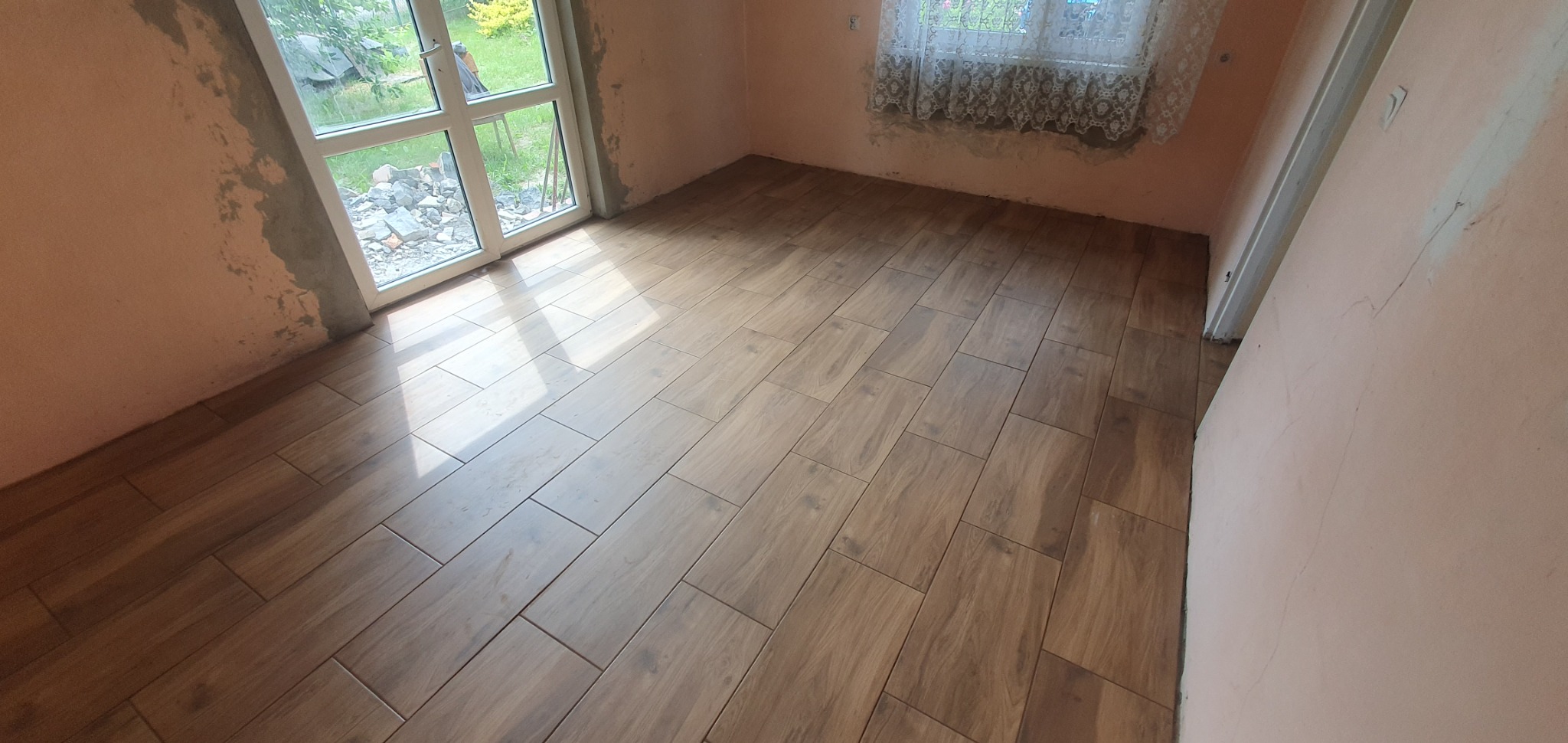 Ułożona podłoga z płytek imitujących drewno w pokoju z surowymi ścianami i oknem na ogród.