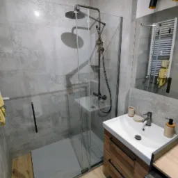 Nowoczesna łazienka z kabiną typu walk-in, szarą armaturą i drewnianą szafką pod umywalką. Widoczne płytki imitujące beton i grzejnik drabinkowy w lustrze.