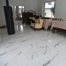 Salon z nową, błyszczącą podłogą z białych płytek imitujących marmur, czarny kominek wolnostojący, meble w stylu vintage i duże okna.