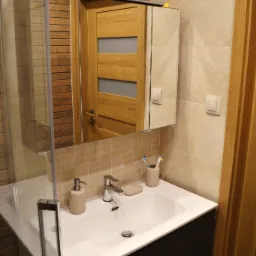 Nowoczesna łazienka z beżowymi płytkami, białą umywalką nablatową na czarnej szafce z połyskiem, lustrem z oświetleniem, kabiną prysznicową z przezroczystego szkła i drewnianymi drzwiami z szybami.