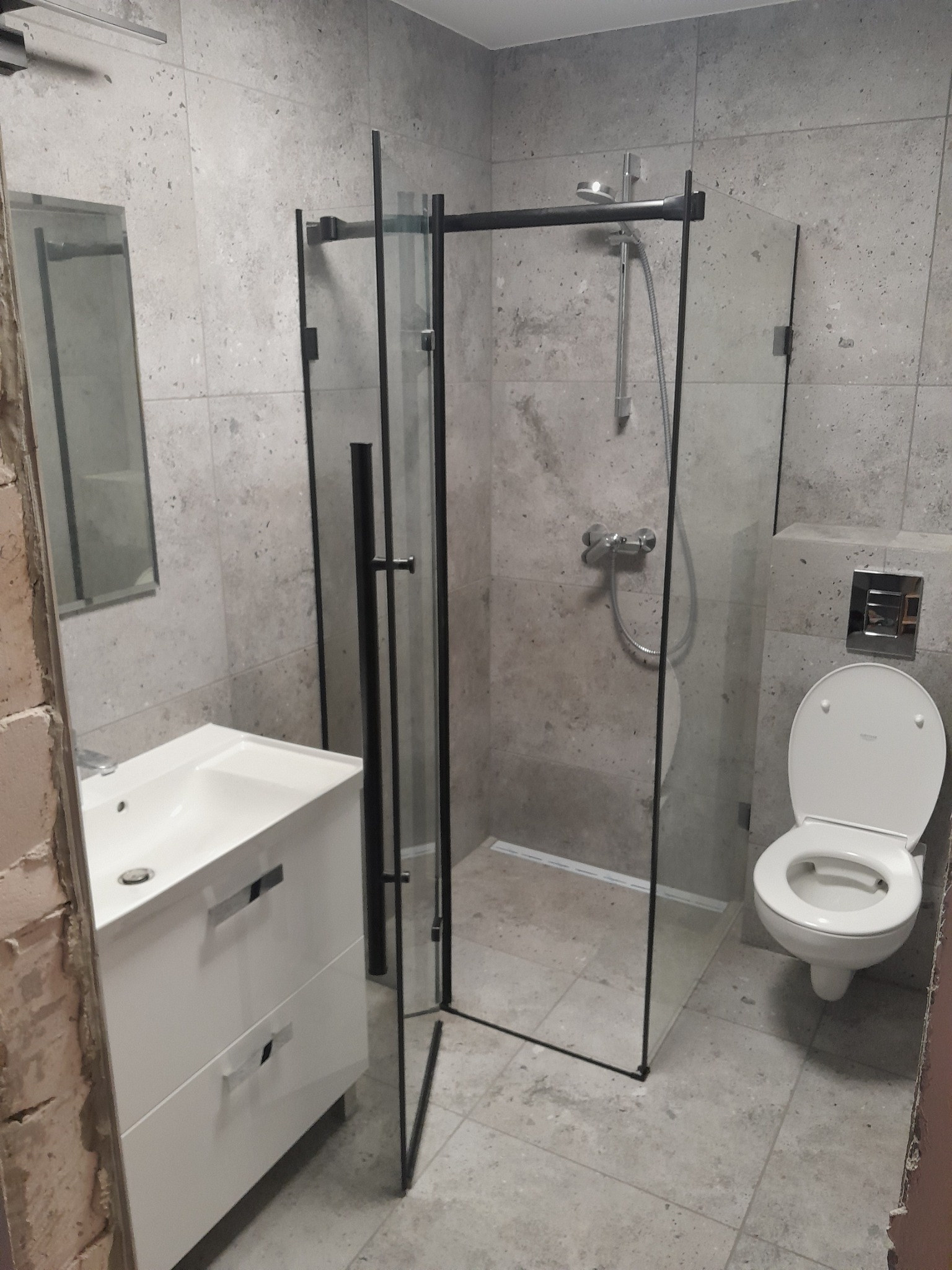 Nowoczesna łazienka z szarymi płytkami imitującymi kamień, kabina prysznicowa typu walk-in z czarnymi profilami, biała umywalka z szafką, lustro oraz toaleta.