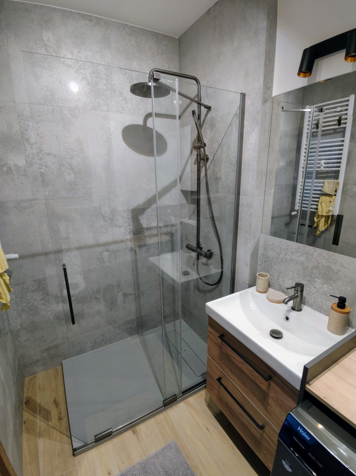 Nowoczesna łazienka z kabiną typu walk-in, szarą armaturą i drewnianą szafką pod umywalką. Widoczne płytki imitujące beton i grzejnik drabinkowy w lustrze.