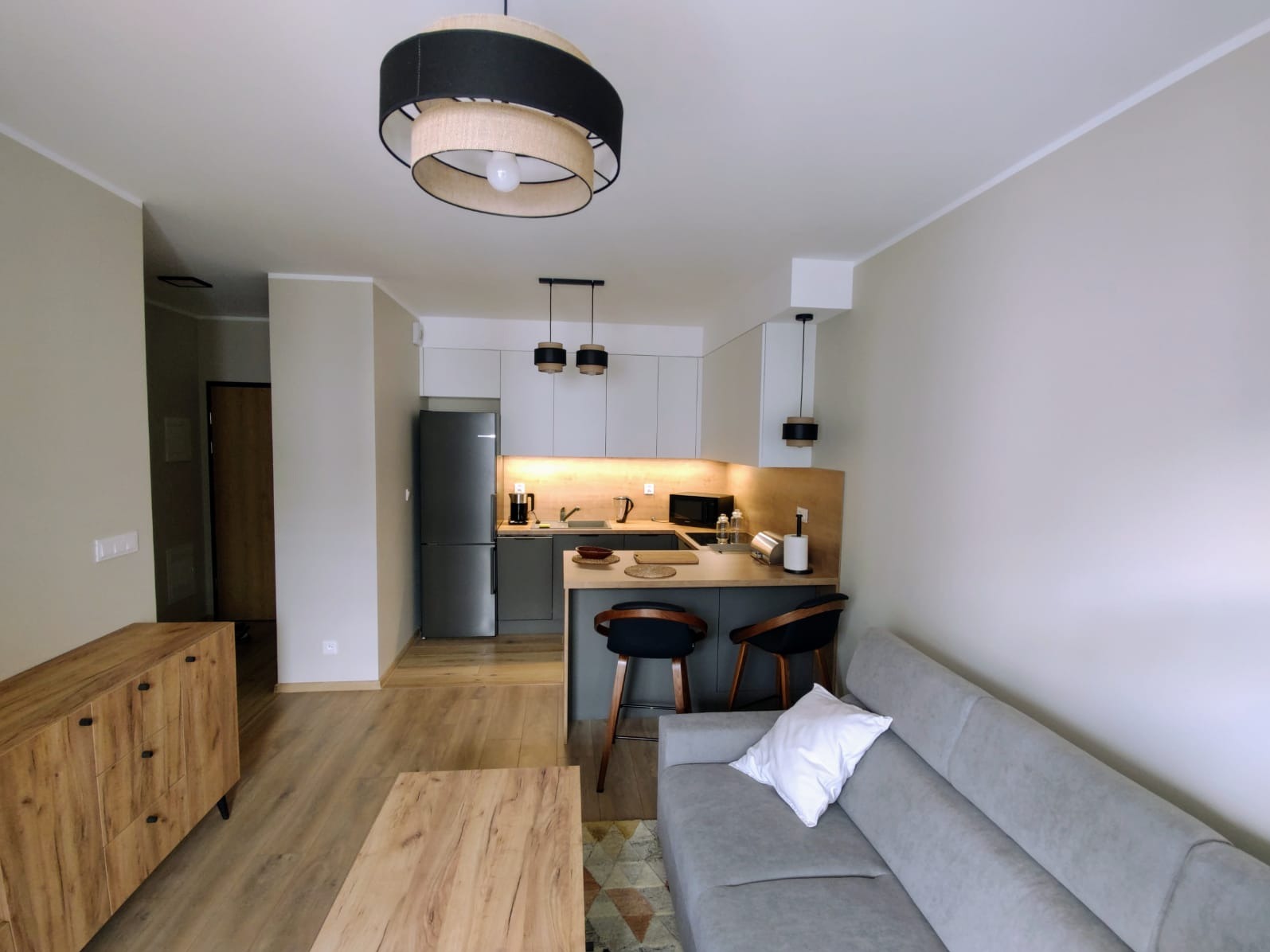 Nowoczesne wykończenie wnętrza mieszkania: jasna kuchnia z barkiem, szara sofa i drewniane meble. Designerskie lampy wiszące dodają elegancji.