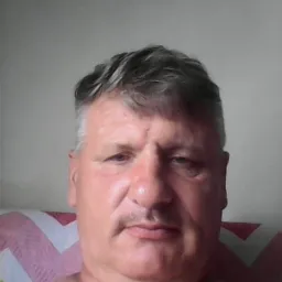 Portret mężczyzny z siwiejącymi włosami i wąsami, na tle jasnej ściany i wzorzystej poduszki, uchwycony w kadrze od ramion w górę.