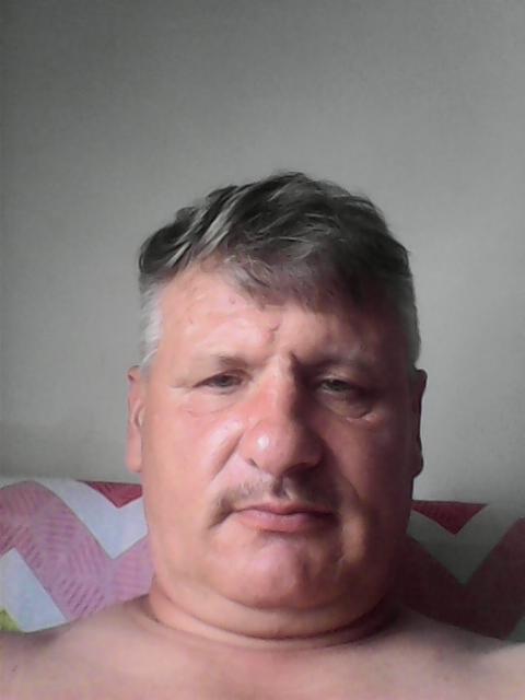 Portret mężczyzny z siwiejącymi włosami i wąsami, na tle jasnej ściany i wzorzystej poduszki, uchwycony w kadrze od ramion w górę.