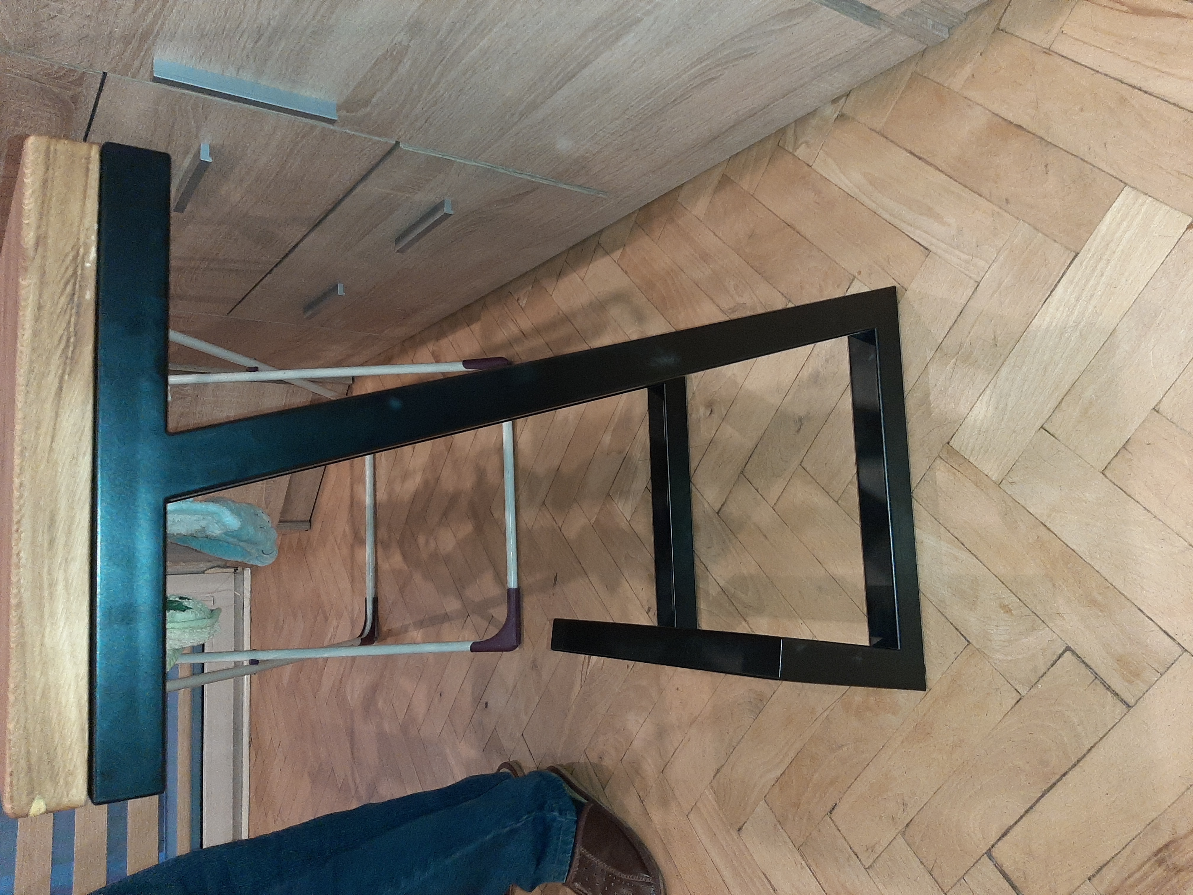 Fragment metalowej konstrukcji, prawdopodobnie ramy, z widocznymi elementami mocującymi i fragmentem podłogi z parkietu w jodełkę, widziane z góry, z butem osoby fotografującej w rogu kadru.