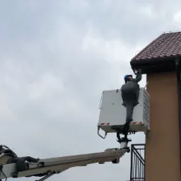 Monter w niebieskim kasku, na podnośniku koszowym, instaluje urządzenie na elewacji domu pod dachem z czerwonej dachówki.