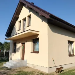 Ocieplony dom jednorodzinny z beżową elewacją, brązowym dachem i oknami, z psem w ogrodzie i wężem ogrodowym przy ścianie.