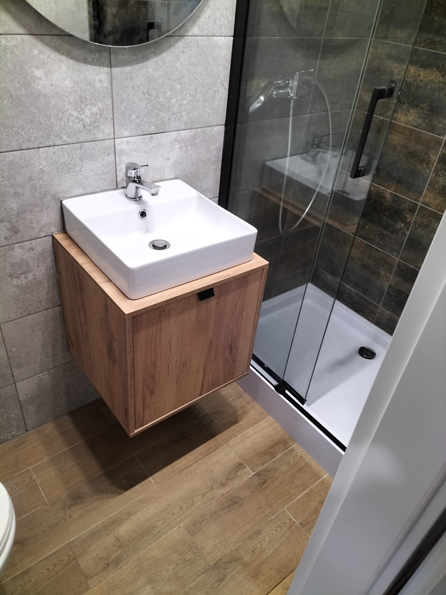 Nowoczesna łazienka: biała umywalka nablatowa na drewnianej szafce, szara płytka ścienna i kabina prysznicowa z czarnymi profilami. Drewnopodobna podłoga.