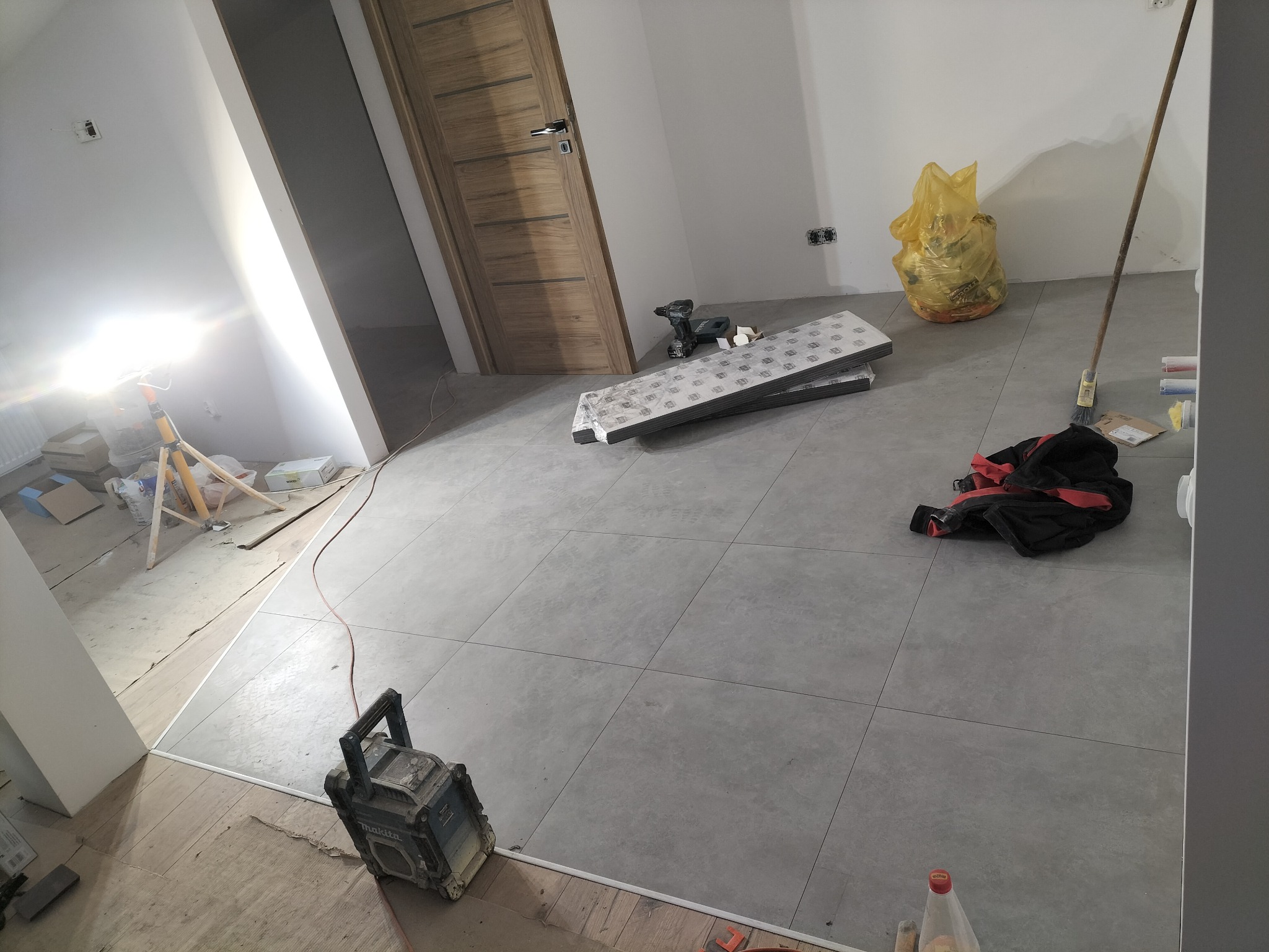 Ułożone szare płytki podłogowe w trakcie remontu. Widoczny sprzęt budowlany, worki z odpadami i fragmenty niezakończonych prac wykończeniowych przy progu.