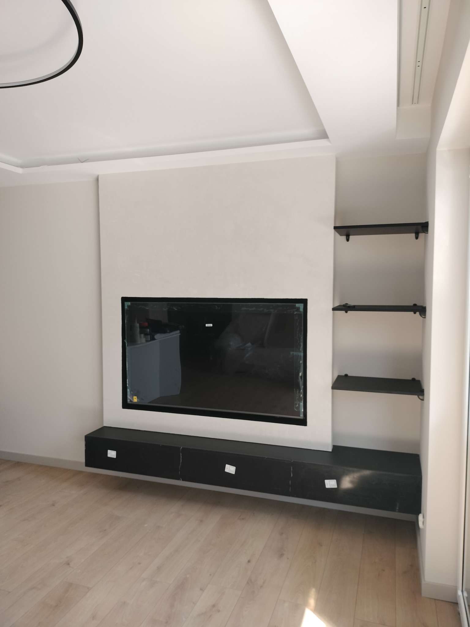 Nowoczesny salon z zabudową TV w kolorze czarnym, minimalistyczne półki na ścianie i jasna podłoga. Całość utrzymana w stonowanej kolorystyce.