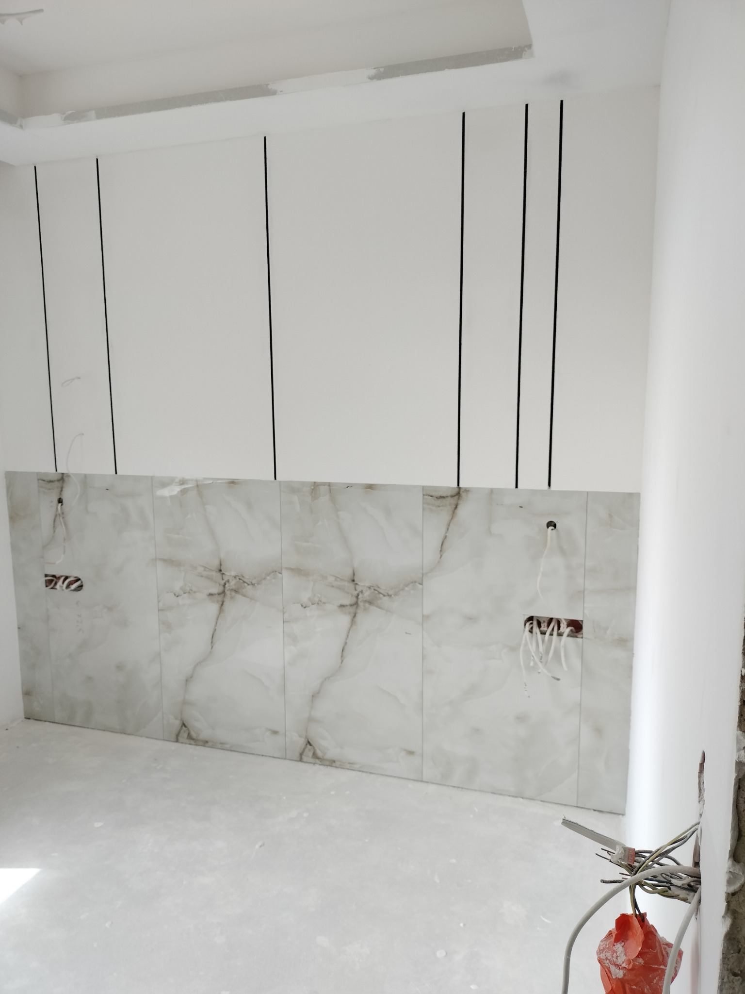 Ściana wyłożona płytkami imitującymi marmur, z widocznymi kablami elektrycznymi i pionowymi czarnymi listwami dekoracyjnymi na białej ścianie. Prace wykończeniowe.