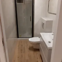 Nowoczesna łazienka z kabiną prysznicową w czarnej ramie, podwieszaną toaletą i białą szafką z umywalką, wykończona płytkami imitującymi marmur i drewnopodobną podłogą.