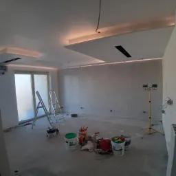 Wnętrze w trakcie prac w Borku Strzelińskim: ściany po nałożeniu gładzi, widoczne drabiny malarskie, wiaderka z farbą i oświetlenie budowlane, przygotowane do dalszych prac wykończeniowych.