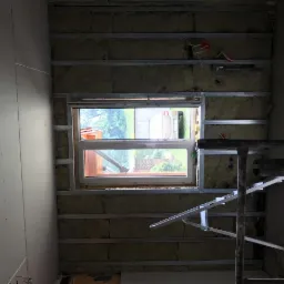 Wnętrze pokoju w trakcie remontu: ściana z oknem, widoczne profile metalowe, izolacja z wełny mineralnej, częściowo zamontowane płyty gipsowo-kartonowe, drabina malarska.