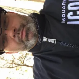 Portret mężczyzny w czapce i okularach, ubrany w kurtkę z napisem 'DSQUARED ICON'. Widoczne drzewo w tle. Zdjęcie obrócone.