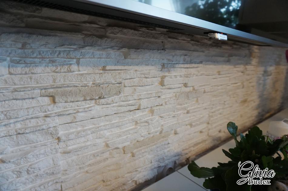 Wykończona ściana z dekoracyjnej, białej cegły pod telewizorem, z widocznym fragmentem rośliny doniczkowej w prawym dolnym rogu.