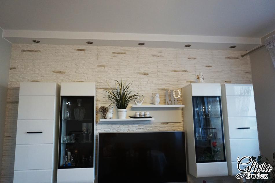 Salon z telewizorem, białymi szafkami z przeszklonymi witrynami i ścianą wyłożoną dekoracyjną cegłą w odcieniach bieli i beżu.