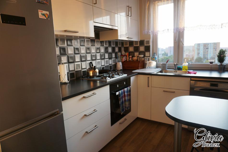 Nowoczesna kuchnia z białymi szafkami, czarnym blatem i mozaiką na ścianie, widok na miasto przez okno, z logo firmy w prawym dolnym rogu.