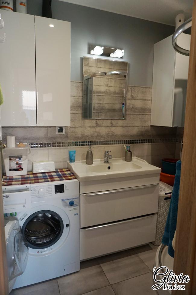 Jasna łazienka z pralką obok szafki umywalkowej, lustro z oświetleniem nad umywalką, płytki imitujące drewno na ścianie.