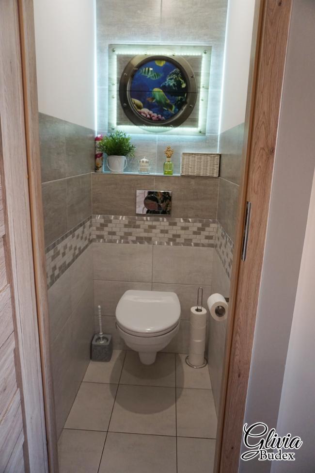 Wykończona toaleta z szarymi płytkami, dekoracyjnym pasem mozaiki, oraz podświetlanym obrazem z motywem morskim nad spłuczką.