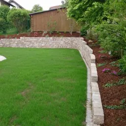 Utrzymany trawnik z niską kamienną ścianą oporową i rabatami wyłożonymi korą, w tle drewniany domek narzędziowy.