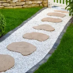 Ścieżka w ogrodzie z nieregularnych kamiennych płyt ułożonych na białym żwirze, obrzeża z ciemnej kostki, trawnik po bokach, mur z kamienia w tle.