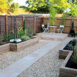 Nowoczesny ogród z podniesionymi rabatami z drewnianych bali, ścieżką z jasnych płyt i żwirowym wypełnieniem, uzupełniony białymi krzesłami i stołem oraz metalowym paleniskiem.