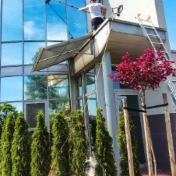 Pracownik myje okna na wysokości nowoczesnego biurowca z użyciem lancy teleskopowej, widoczna drabina i dekoracyjne drzewa.