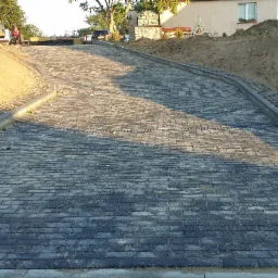 Ukończony podjazd z kostki brukowej w kolorze grafitowym, wykończony betonowymi obrzeżami i kratką trawnikową, z widoczną studzienką kanalizacyjną i przygotowanym terenem pod dalsze prace ziemne...