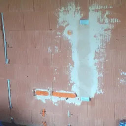 Ściana z cegły z zamontowanymi puszkami elektrycznymi i przewodami, przygotowana pod instalację. Widoczne przewody w peszlach i rury instalacyjne na podłodze.