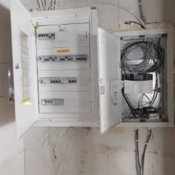 Otwarta szafka elektryczna z bezpiecznikami i listwami zaciskowymi po lewej stronie oraz centralą alarmową z okablowaniem po prawej stronie, widoczne przewody w ścianie.