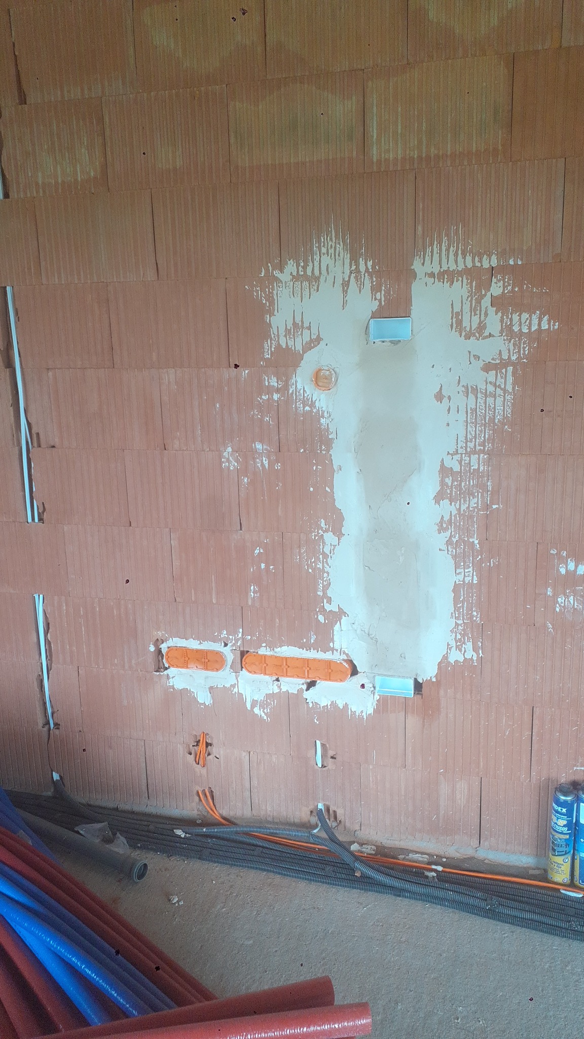 Ściana z cegły z zamontowanymi puszkami elektrycznymi i przewodami, przygotowana pod instalację. Widoczne przewody w peszlach i rury instalacyjne na podłodze.