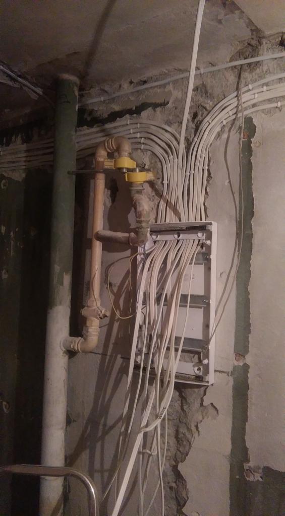 Białe przewody elektryczne wychodzące z puszki na surowej ścianie z widocznymi rurami i instalacjami hydraulicznymi w tle.